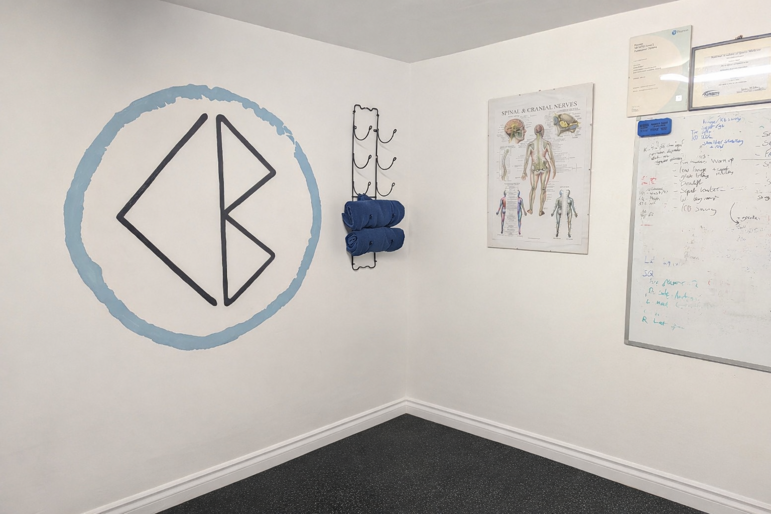 Corner of a physiotherapy clinic room featuring wall logo, spinal nerve anatomy poster, mounted towel rack with blue towels, and whiteboard with handwritten notes.